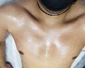 A person lying down with an oily chest and wearing a black face mask and a beaded necklace.